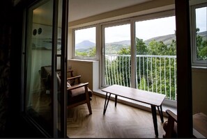 Un balcón con mesa y sillas de madera, un ventanal de vidrio y vistas a colinas verdes.