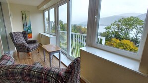 Un balcó amb vista a un llac i a muntanyes, moblat amb un sofà escocès, una cadira i una taula petita.