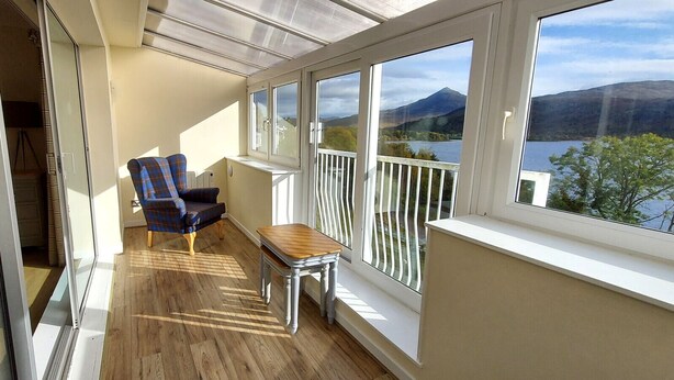 A balcony with a wooden floor, a blue plaid chair, a small wooden table, and a view of a lake and mountains. - Loch Rannoch Highland Club, Pine Martin Lodge 2 (Kinloch Rannoch)