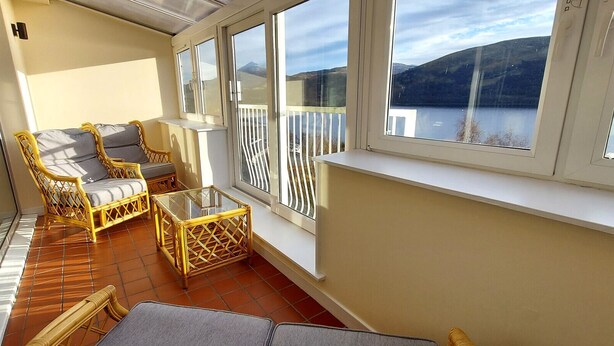Un balcón con muebles de mimbre, una mesa de vidrio y vistas a un lago y montañas.