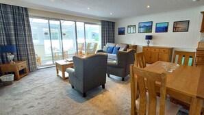 A spacious living room with a sofa, armchairs, a dining table, and a view through large windows.