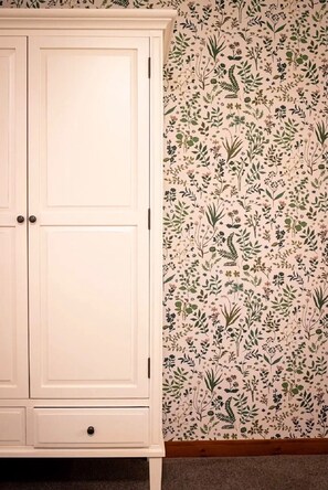 Een witte garderobe met twee deuren en een lade, naast een muur met een bloemen behangpatroon.