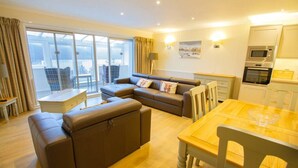 A modern living room with a sofa, ottoman, and dining table. There is a kitchen area with appliances and a large window with curtains. - Rannoch Highland Club, Pine Martin Lodge 6 (Kinloch Rannoch)