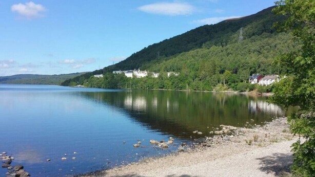 A calm lake with a rocky shoreline and a forested hillside in the background. - Rannoch Highland Club, Pine Martin Lodge 6 (Kinloch Rannoch)
