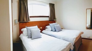 A hotel room with two single beds, each with white bedding and grey pillows. There is a wooden headboard, a window with curtains, and a mirror on the wall. - Loch Rannoch Highland Lodges Apartment 13 (Kinloch Rannoch)