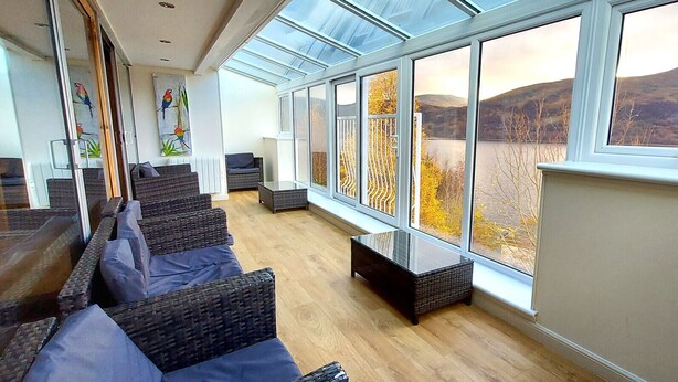 A modern interior with a glass ceiling, wicker furniture, and a view of a lake and hills. - Loch Rannoch Highland Lodge 7 (Kinloch Rannoch)