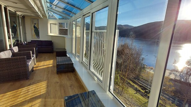 Ein modernes Interieur mit Glasdecke, Korbmöbeln und Blick auf einen See und Hügel.