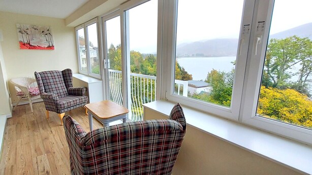 A balcony with a view of a lake and trees, furnished with plaid chairs and a small table.