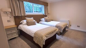 A bedroom with two beds, a window with curtains, a radiator, and a bedside table.