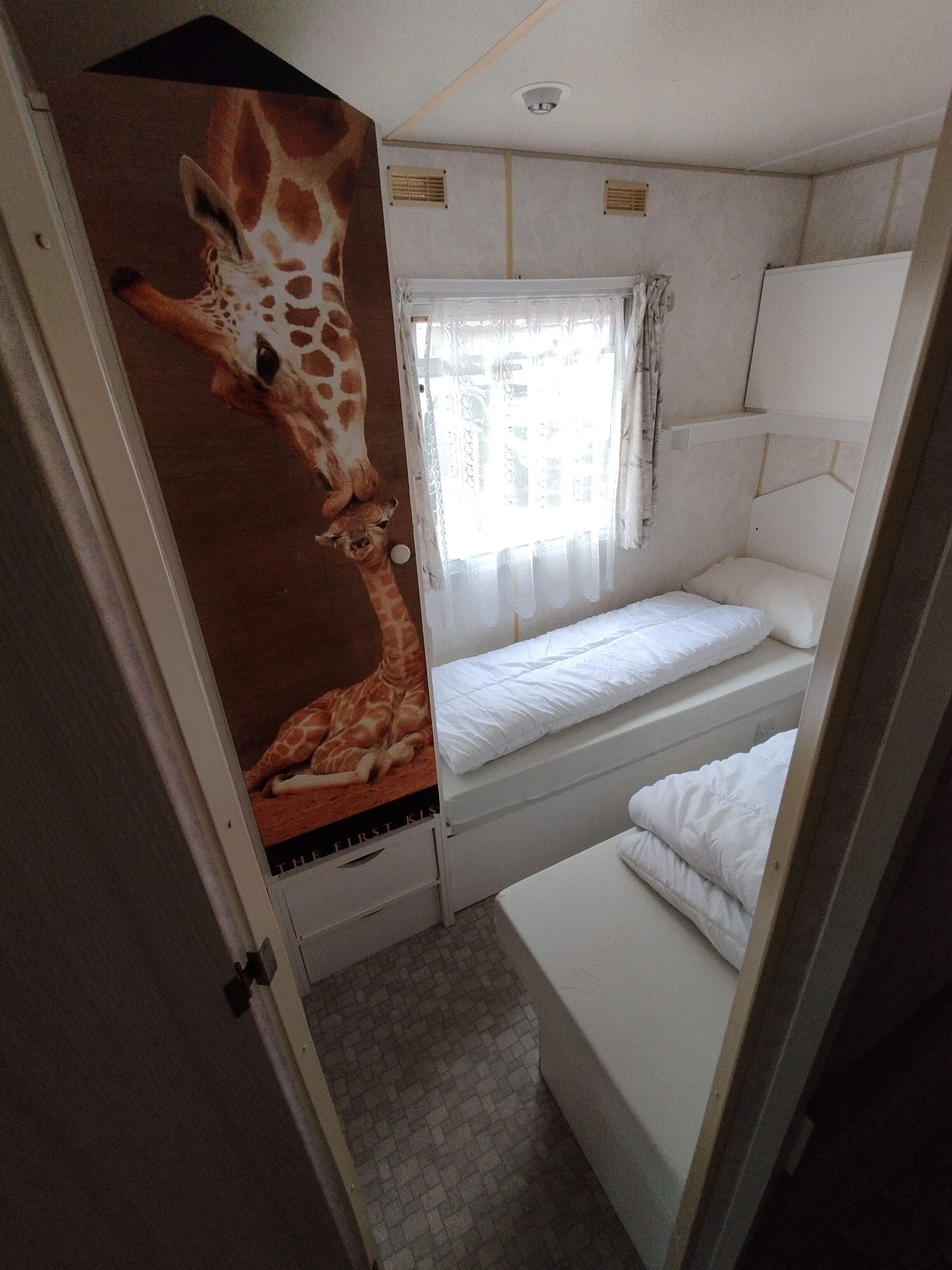 Ein kleines Zimmer mit Etagenbett, einem Fenster mit durchsichtigen Vorhängen und einem Giraffenplakat an der Wand.