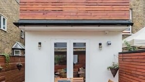 Un patio con una mesa y sillas de madera, un pequeño edificio blanco con una puerta y un muro de ladrillo.