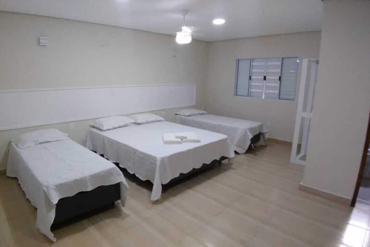 A room with three single beds, each with a white bedspread and matching pillows. The room has a tiled floor and a white headboard against the wall.