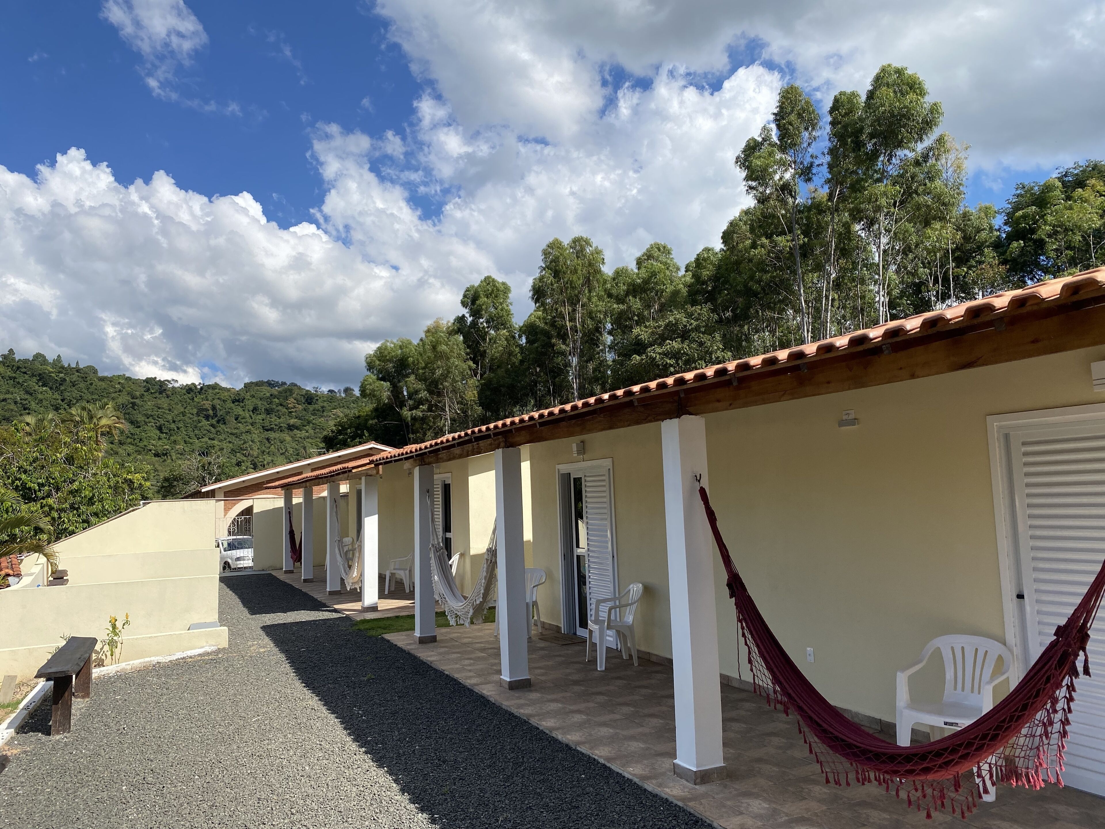 A row of single-story buildings with white doors and windows, a paved walkway, and a view of a forested hill.