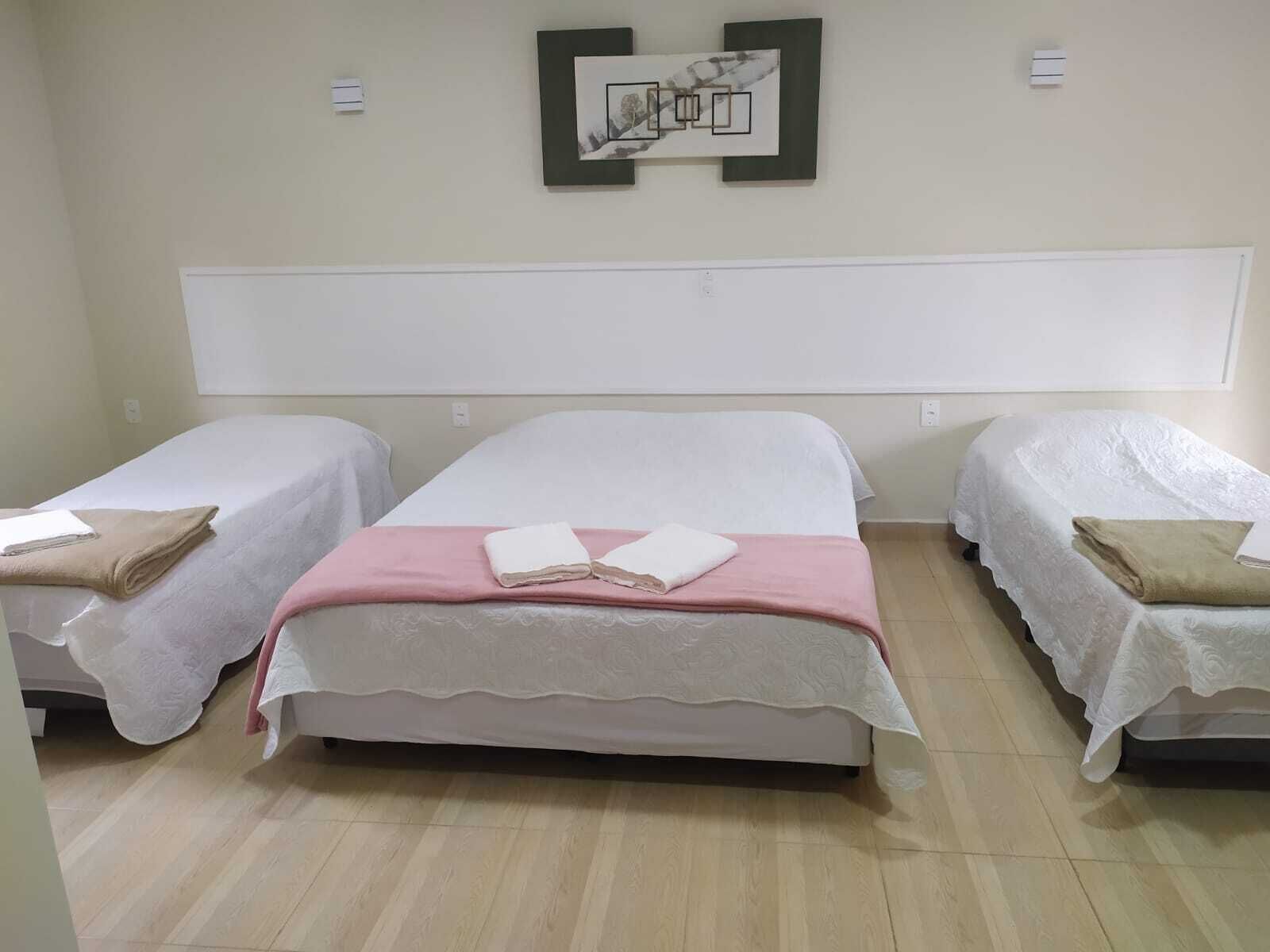 Habitación de hotel con tres camas, cada una con cubrecama blanco y una manta rosa en la cama central. Hay dos unidades de aire acondicionado fijadas a la pared y un cuadro enmarcado sobre las camas.