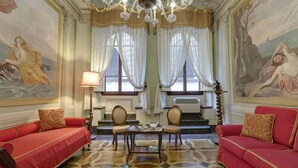 A room with a red sofa, a table with a lamp, and two chairs. There are two windows with curtains, a chandelier, and a painting on the wall.