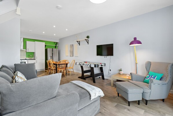 A modern living room with a grey sofa, a houndstooth armchair, a small table, and a dining area with a green accent wall.