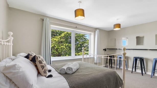 Una cama bien hecha con almohadas y una toalla enrollada, una ventana con cortinas, y una lámpara colgante.