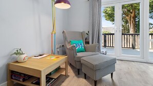 Una habitación acogedora con un sillón a cuadros de pata de gallo, una mesita de madera con juegos de mesa, una lámpara de pie y vistas al exterior a través de una puerta corredera de vidrio.
