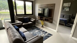 Una sala de estar moderna con un sofá grande, un televisor de pantalla plana montado en la pared y vistas al exterior a través de amplias ventanas.