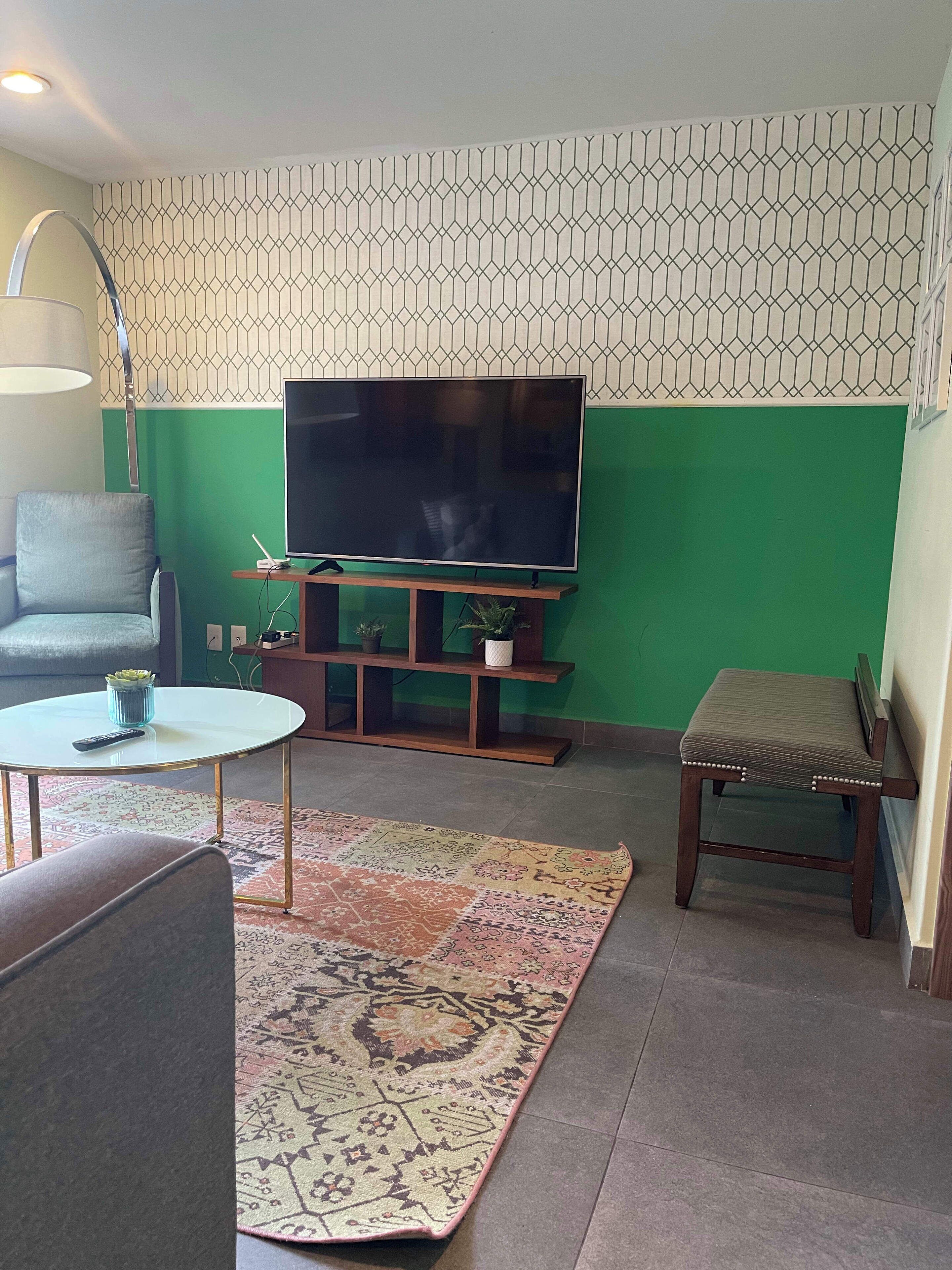 A living room with a green wall, a flat-screen TV on a wooden stand, a sofa, a small round table with a remote, a patterned rug, and a bench.