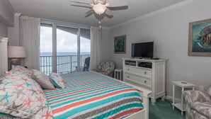 A hotel room with a bed, a ceiling fan, a TV, a dresser, and a view of the ocean.