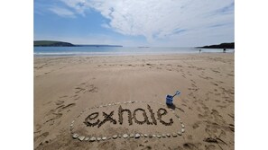 A beach with footprints and the word "exhale" written in the sand, a blue bucket, and a distant sailboat.