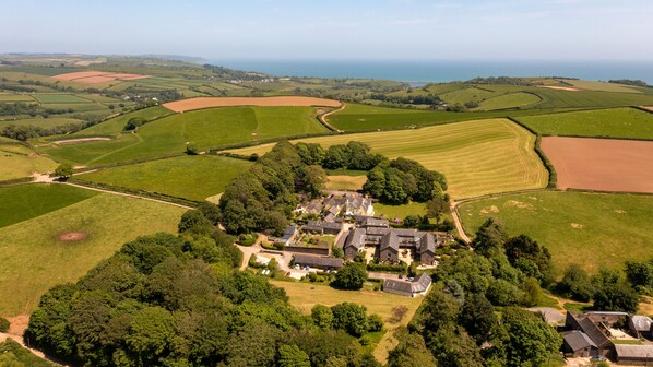 An aerial view of a rural estate with multiple buildings, surrounded by expansive fields and a coastline in the background.