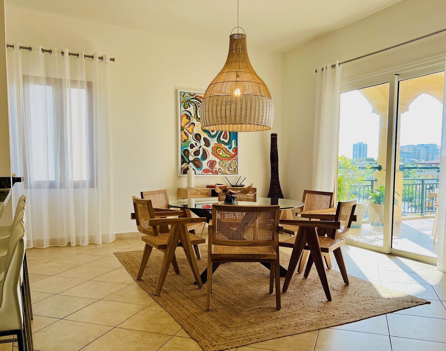 A dining area with a round table, six wooden chairs, a woven pendant light, and a large abstract painting on the wall.