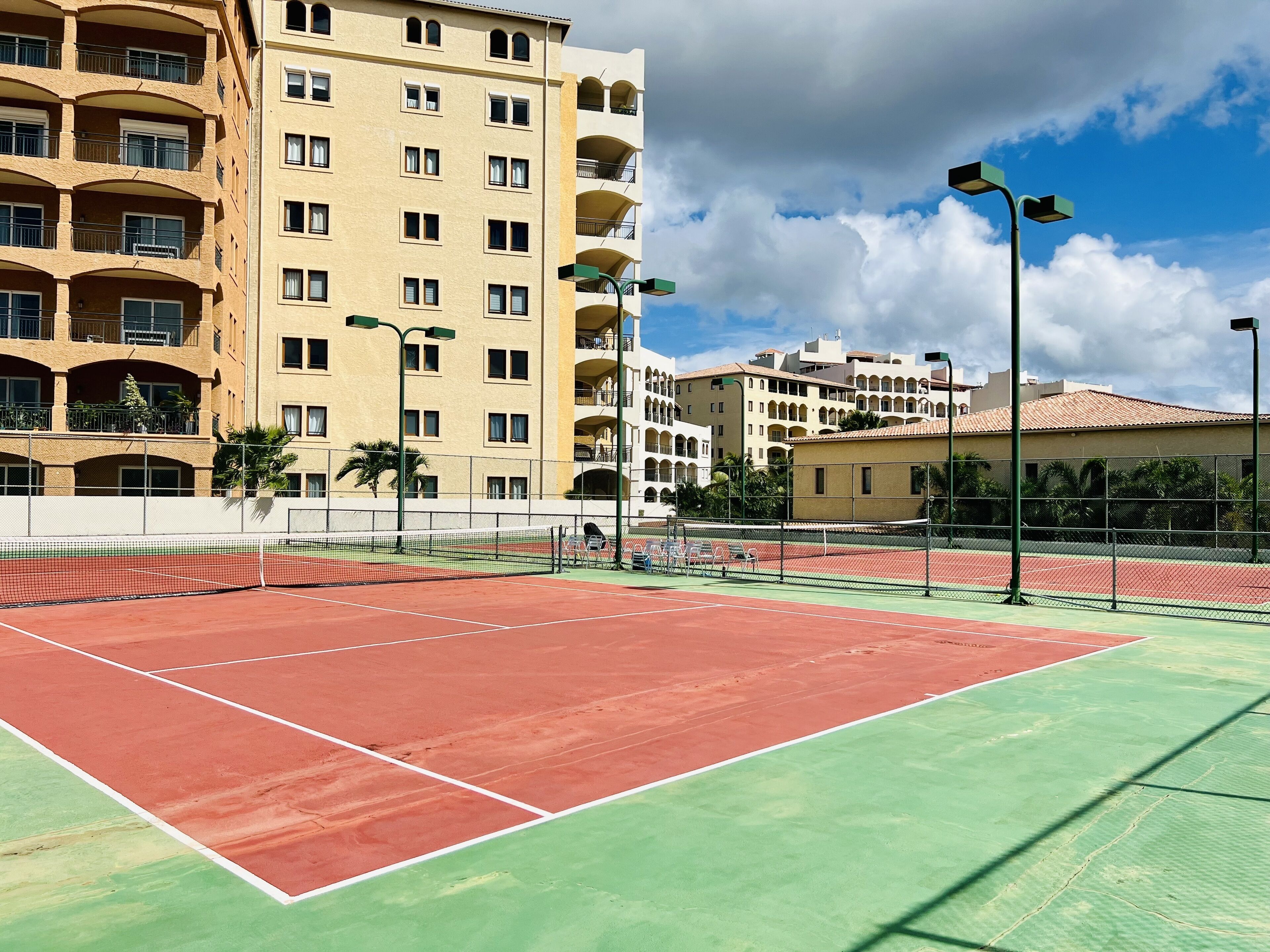 A tennis court with a net, surrounded by a fence, and adjacent to a multi-story building with balconies.