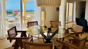 A dining area with a glass table surrounded by wicker chairs, a large woven pendant light, and a view of the sea and buildings through large glass doors. - Rendez-vous in St-Martin - Tropical Waves (Sint Maarten)