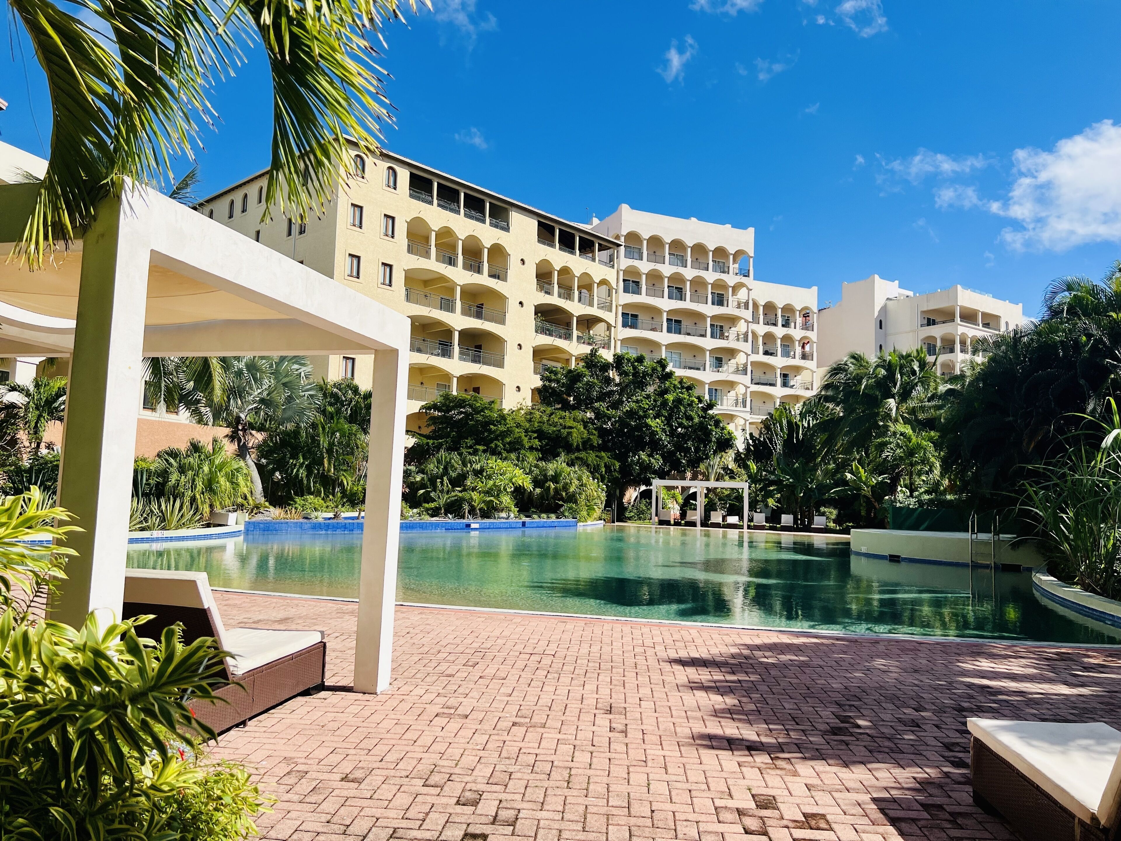 Une piscine d’un complexe hôtelier, avec un ciel bleu azur, des palmiers et un immeuble à plusieurs étages doté de balcons.