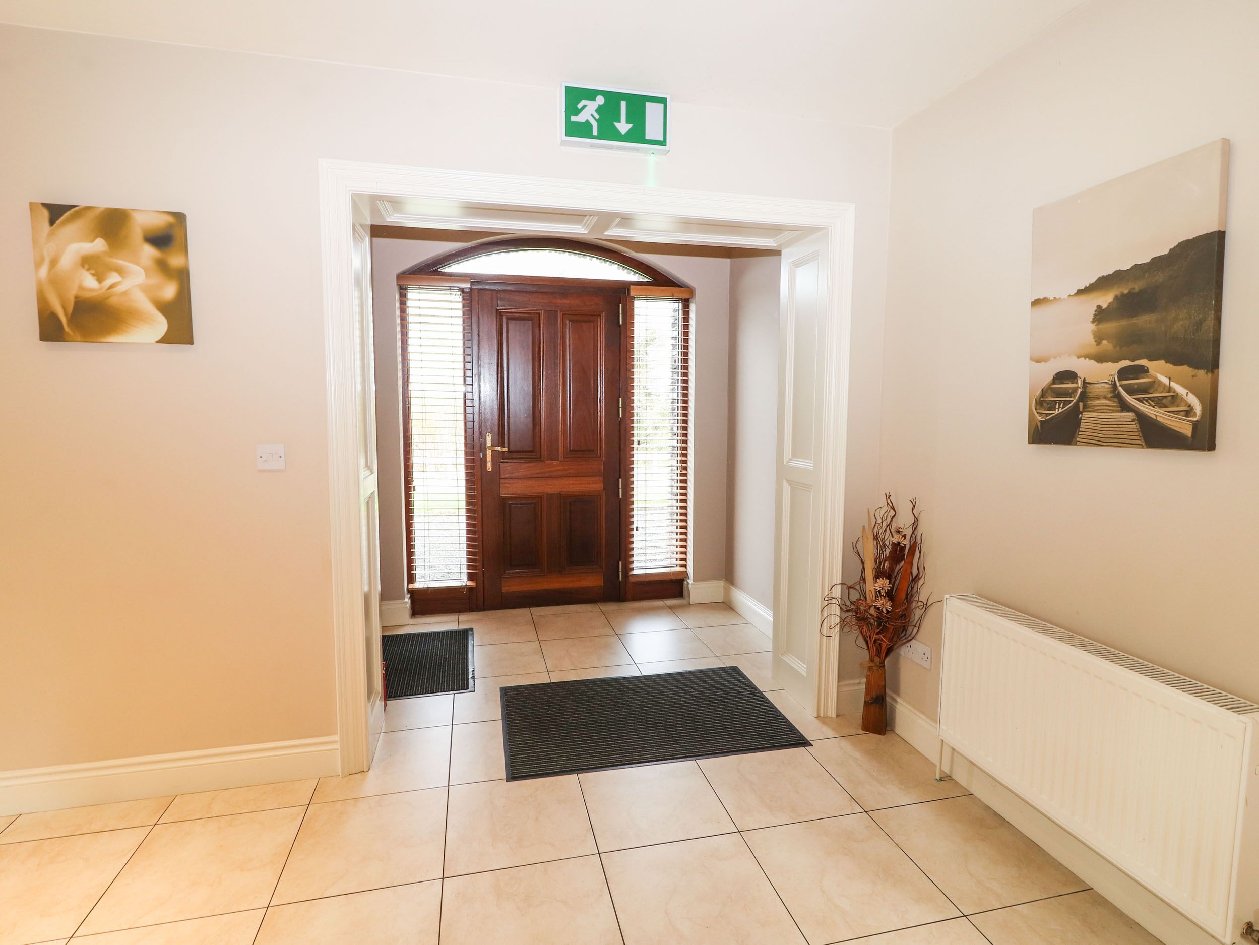 A hotel entrance with a wooden door, a green emergency exit sign, a doormat, a radiator, and two paintings on the walls.