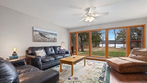 A living room with a leather sofa, a wooden coffee table, and a large window with a view of trees and a lake. - Ninnie & Papaw's Paradise Cove on Beaver Lake (Springdale)