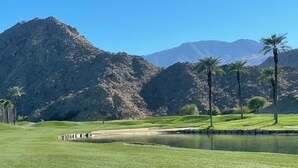 Un campo de golf con un estanque, palmeras y montañas al fondo.