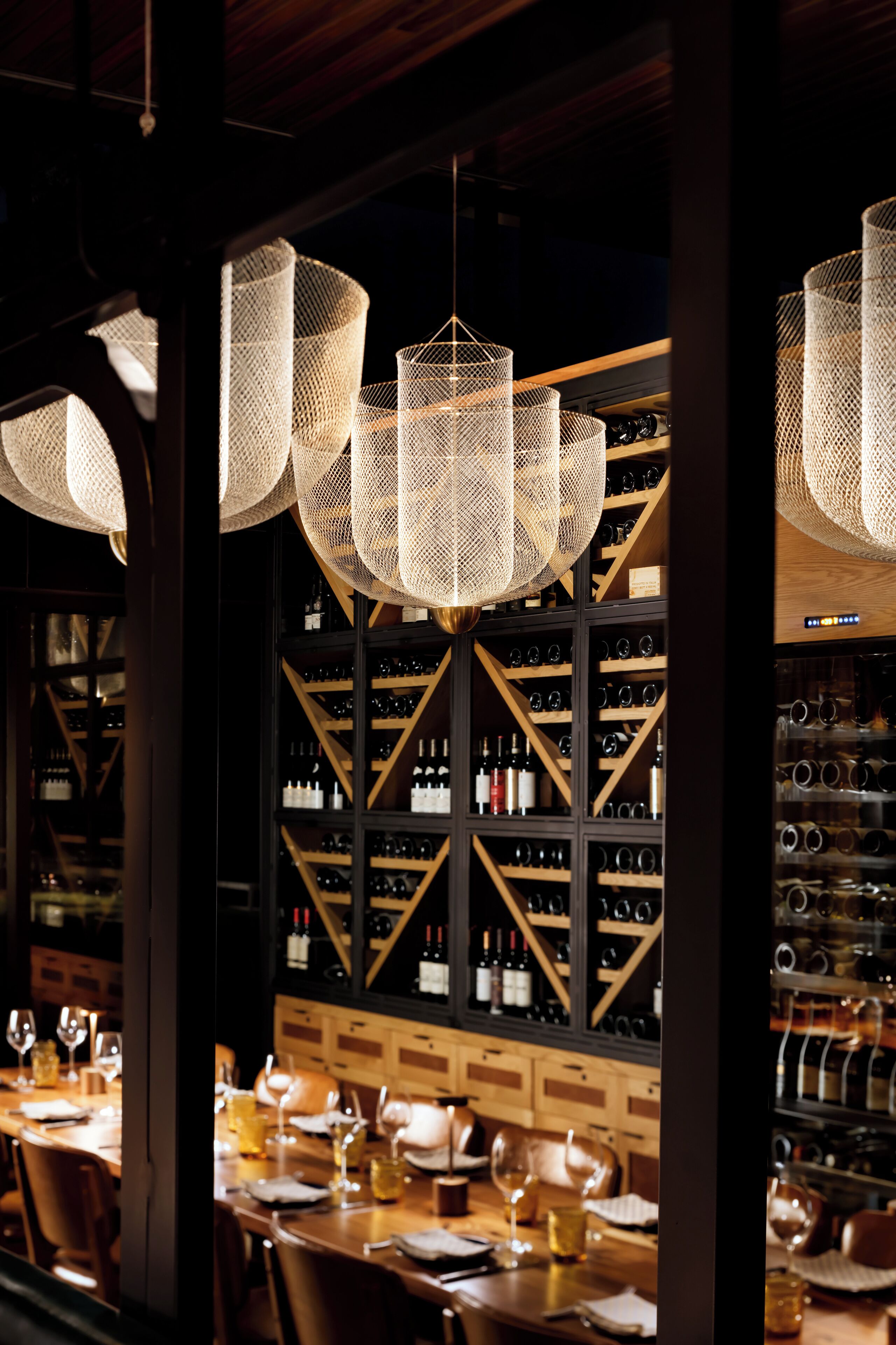 A restaurant with a long table set for dining, a wine rack in the background, and hanging woven light fixtures.