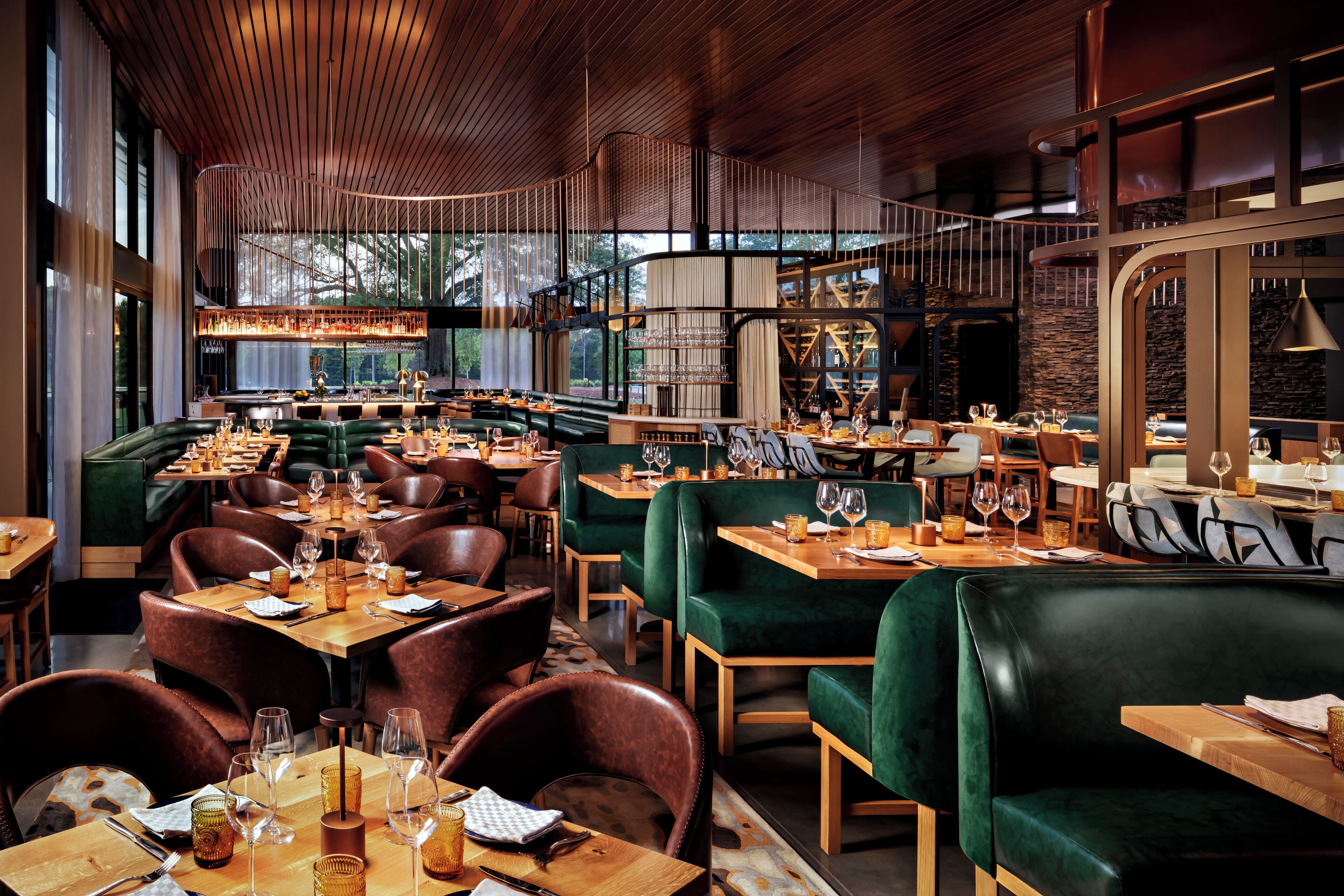 A restaurant with wooden tables, green upholstered booths, and a high ceiling with exposed beams.