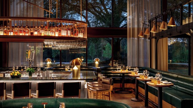 Un restaurante con zona de bar, asientos tapizados en verde y vistas a árboles a través de amplios ventanales.