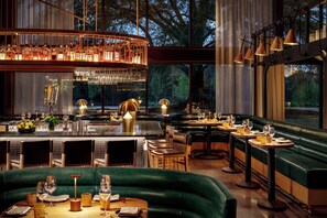 A restaurant with a bar area, green upholstered seating, and a view of trees through large windows. - Hotel Hartness (Greenville)