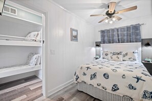 Une chambre avec un lit superposé, un lit simple avec une couverture à motifs d’étoiles de mer et de coquillages, un ventilateur de plafond et une fenêtre avec des rideaux.