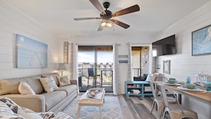A living room with a sofa, coffee table, and a dining area with chairs and a table. There is a ceiling fan, a TV mounted on the wall, and a view of the outside through a sliding door.