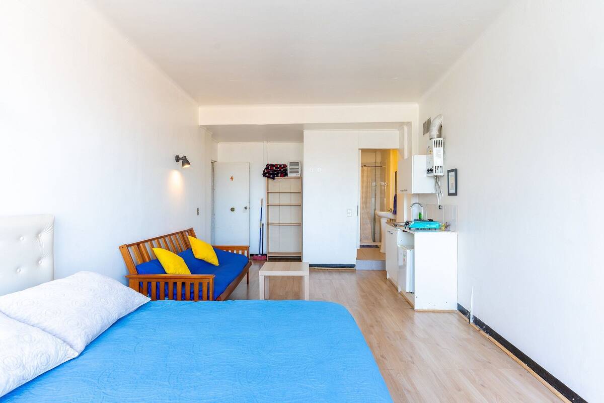 Ein Schlafzimmer mit einem Bett, einer hölzernen Bank mit Kissen, einem kleinen Tisch und einer Küchenzeile mit Spüle und Schränken.