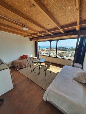 A room with a bed, a small table with chairs, a sofa, and a view of the city and sea through large windows. - Bocamar Apart Hotel  (Concon)