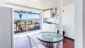 Un balcón con mesa y sillas de vidrio, una zona de cocina blanca y vistas a la ciudad y al océano.