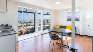 Una habitación amplia con un gran ventanal corredera que da a un balcón, una mesa redonda con sillas y una pequeña cocina con lavamanos.