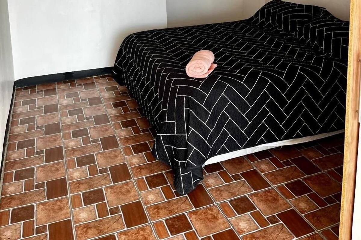 Ein kleines, ordentlich eingerichtetes Schlafzimmer mit gefliestem Boden, einem Bett mit schwarz-weiß gestreifter Bettdecke und einem Holzregal darüber.