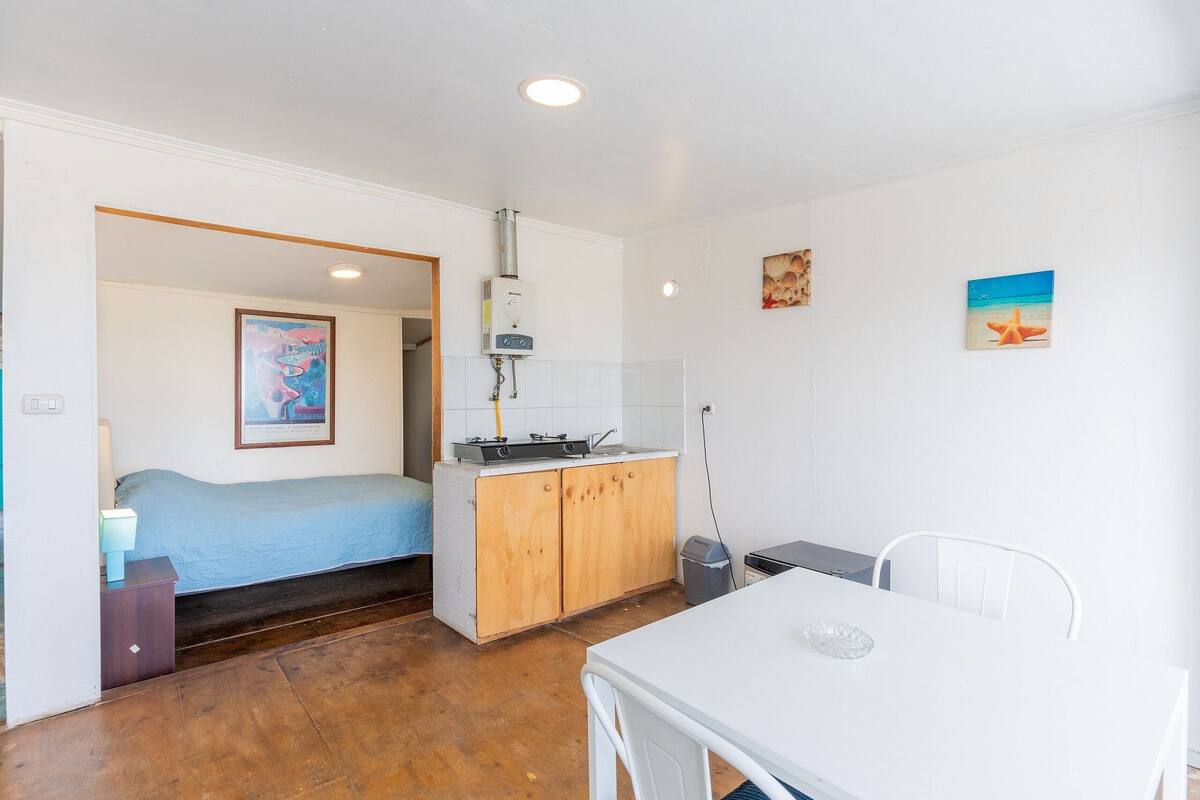 Ein kompakter Raum mit einem Bett, einer kleinen Küchenzeile, einem Esstisch und einer weißen Decke.