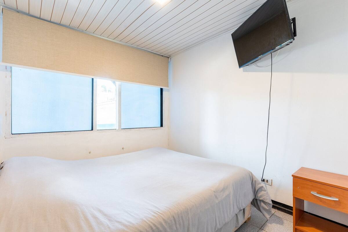 Ein Schlafzimmer mit einem Bett, einem Nachttisch aus Holz, einem an der Wand montierten Fernseher und zwei Fenstern.