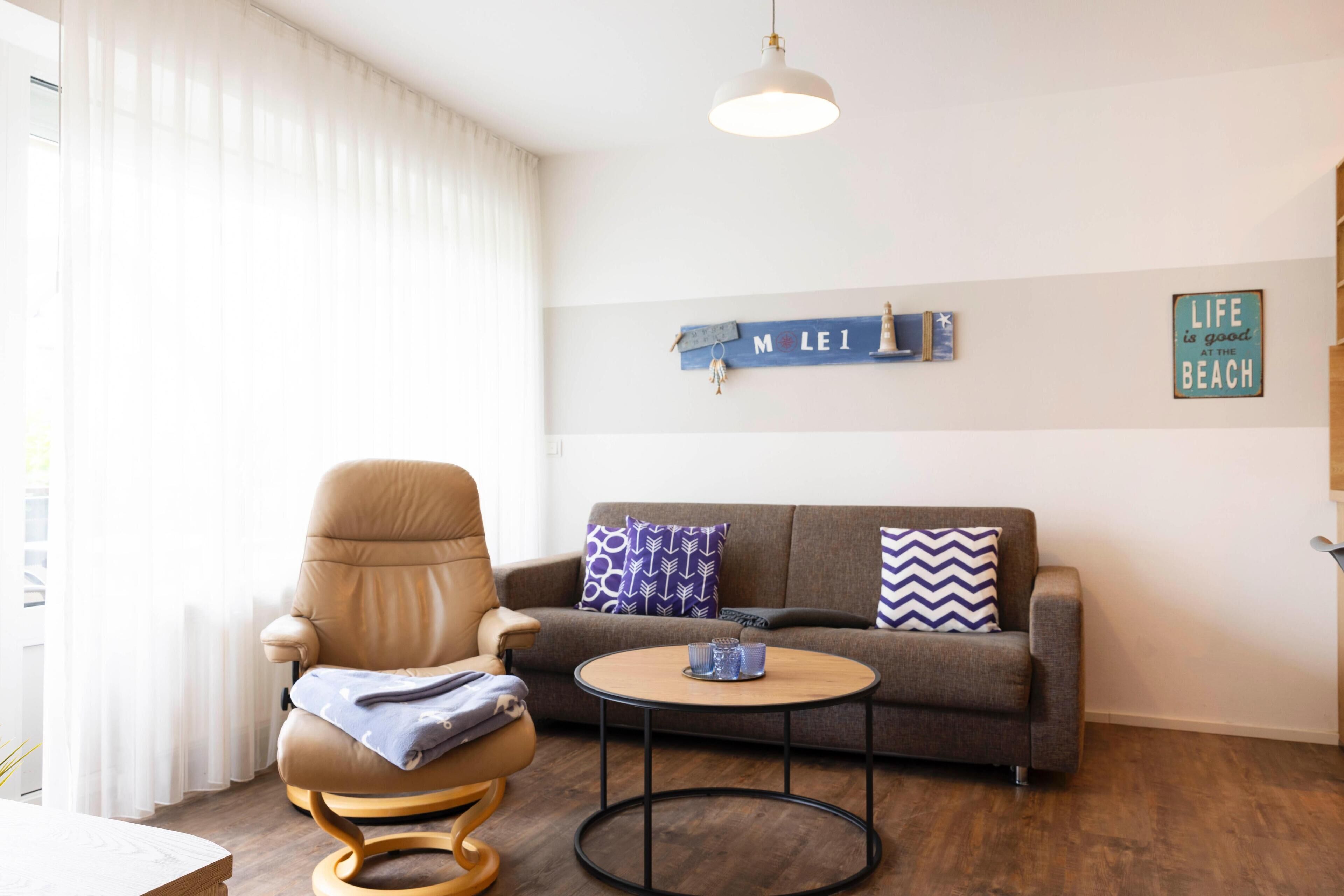 Un salon avec un fauteuil en cuir marron, un canapé gris orné de coussins à motifs, une table basse ronde et un panneau mural sur lequel on peut lire « MOLÉI ».