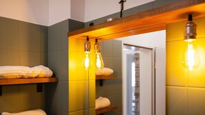 Une salle de bain avec des étagères en bois où sont rangées les serviettes, un miroir avec un cadre en bois et des ampoules apparentes.