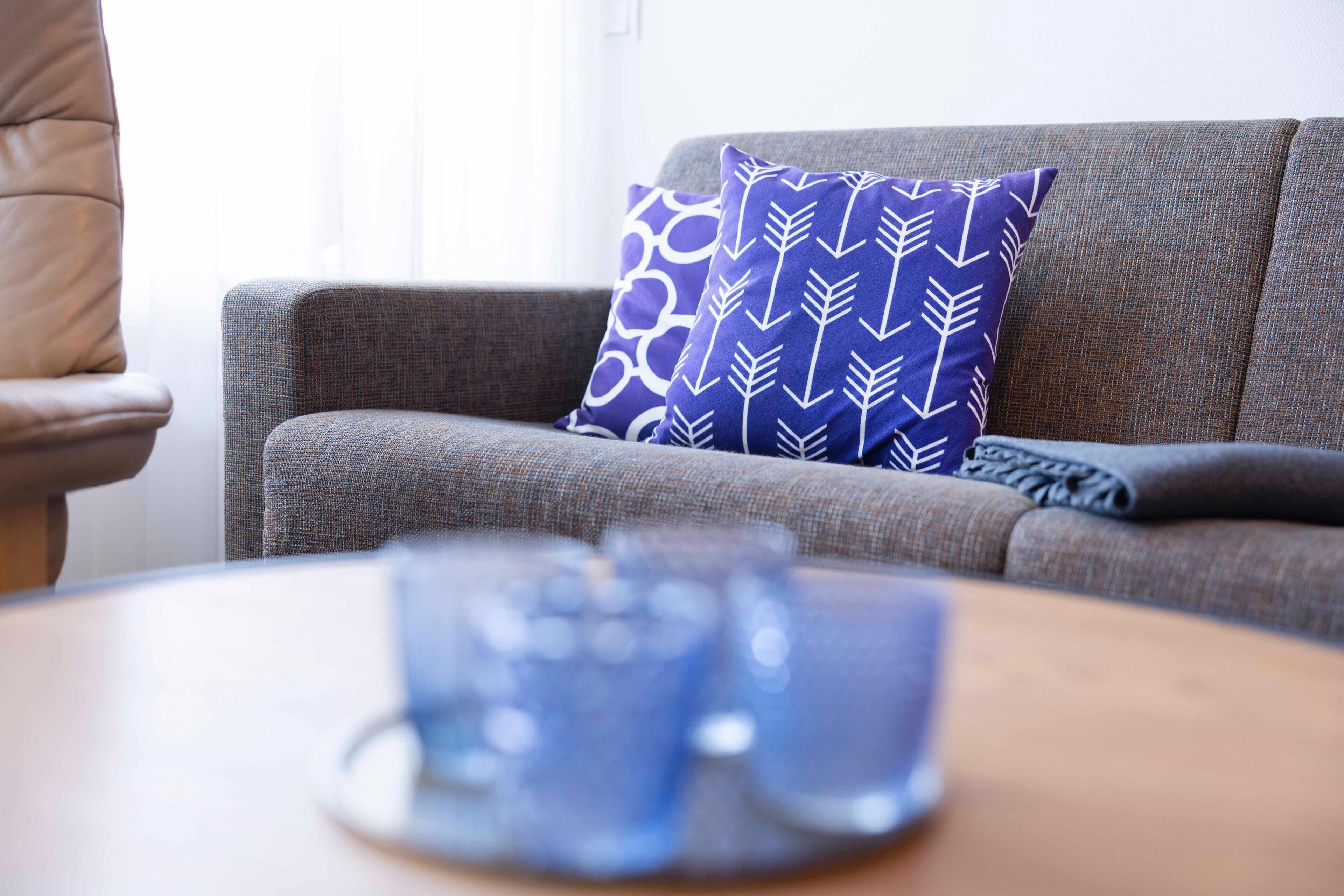 Un sofá gris con un cojín estampado, un control remoto y una mesa de centro con una taza azul.